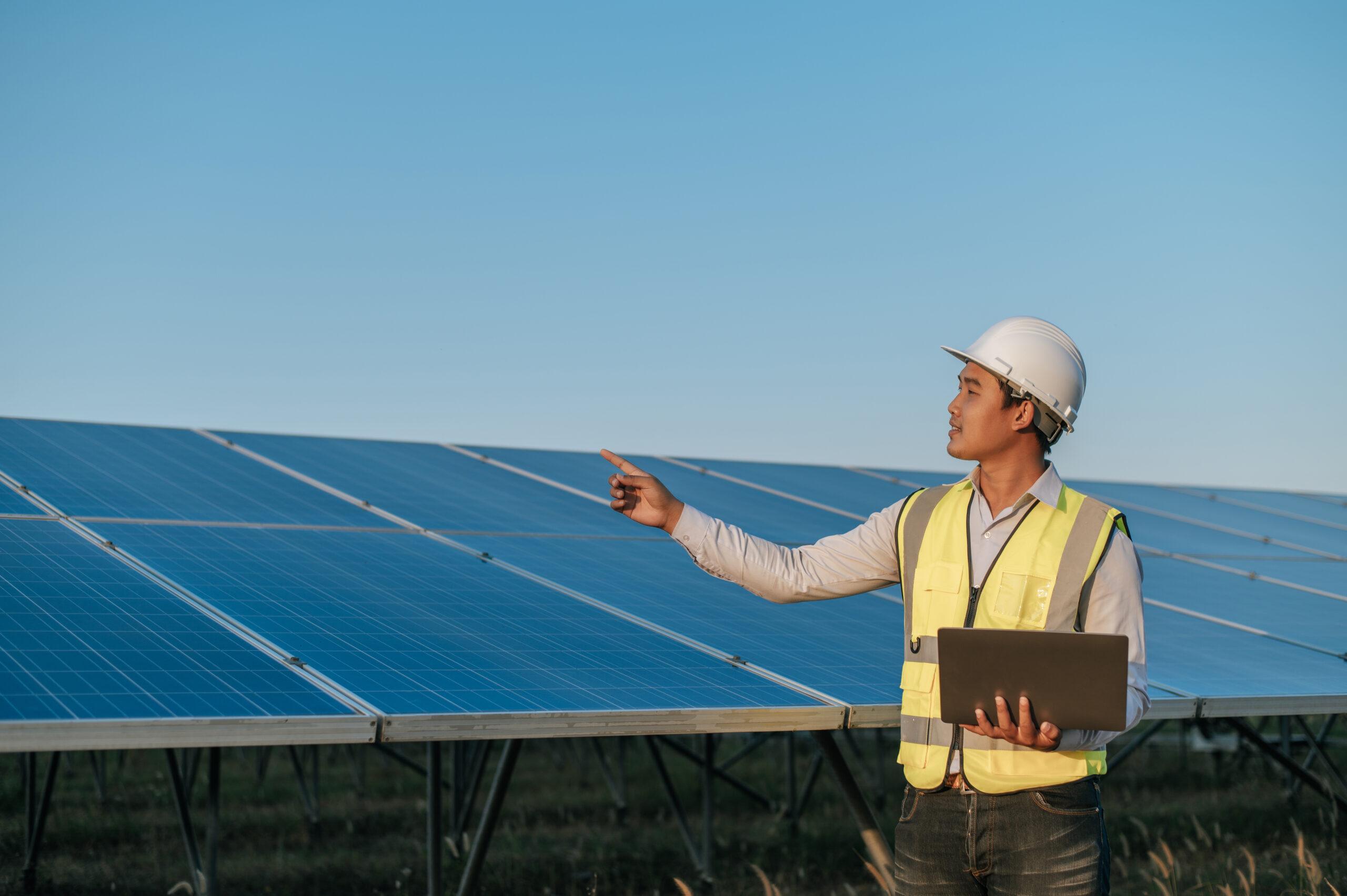 Young Asian Inspector Engineer man use laptop computer working at solar farm, Technician, supervisor male in white helmet Checking operation of sun and photovoltaic solar panel in station, copy space
